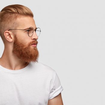 Profile shot of handsome male with trendy hairdo and beard, looks aside with serious expression, has thick red beard, wears round glasses, focused into distance, isolated over white background