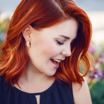 Closeup portrait of smiling laughing flirty middle aged white caucasian woman with waved curly red hair in black dress screaming outside in park, beauty fashion lifestyle concept